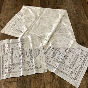 May include: Three white linen tablecloths with an intricate embroidered pattern. The tablecloths are folded and laid out on a wooden floor.