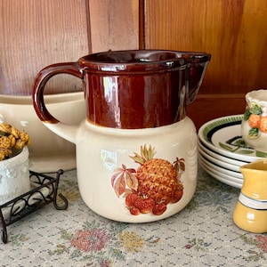 May include: A vintage ceramic pitcher with a brown top and a cream-colored base. The base features a pineapple and strawberry design. A small yellow pitcher and a stack of plates are also visible.