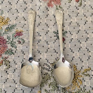May include: Two silver-toned metal spoons with ornate handles. The left spoon features a square-shaped bowl, while the right spoon has a traditional oval-shaped bowl. Both handles are engraved with a crown emblem and decorative detailing.