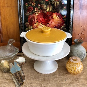 May include: A white ceramic casserole dish with a yellow lid. The dish is sitting on a white pedestal cake stand. The dish is on a burlap surface with a glass chicken dish, silver serving spoons, and a decorative glass vase.