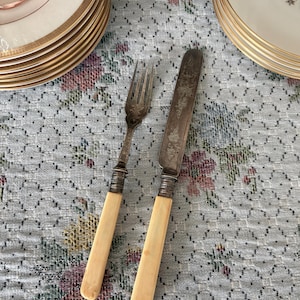May include: A vintage fork and knife set with cream-colored handles and ornate silver detailing. The fork has four tines, and the knife features a decorative floral pattern. The set is placed on a floral patterned tablecloth, with stacks of plates in the background.