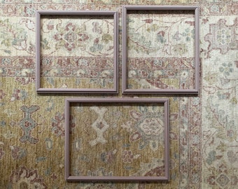 May include: Three empty picture frames with a light pink wood finish are arranged in a triangular pattern on a patterned rug. The rug has a beige background with a floral design in shades of brown, red, and blue.