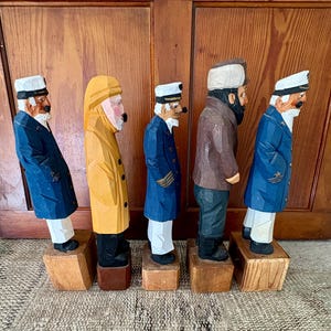 May include: Five carved wooden sailor figurines. The figures are dressed in various coats and hats, with white beards and mustaches. The color palette includes blue, yellow, brown, and white. Each figure stands on a small wooden block.