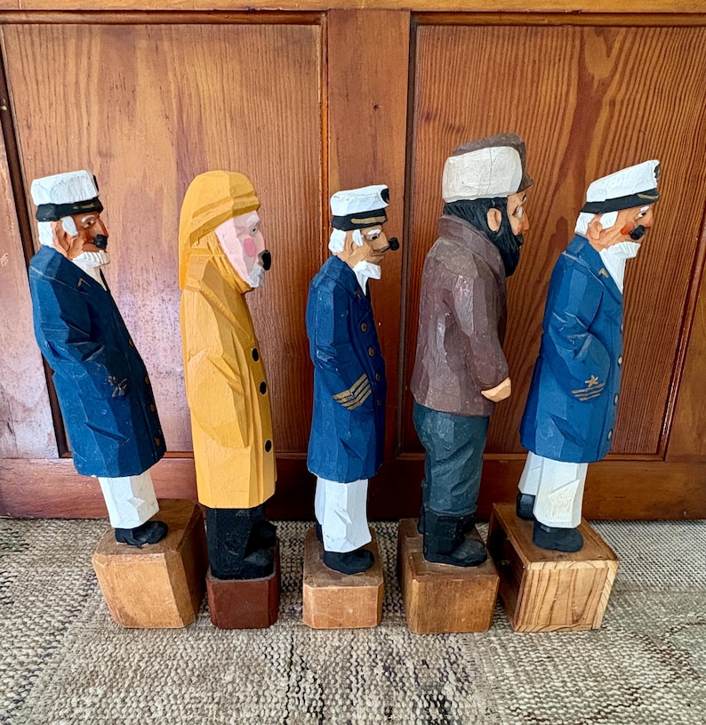 May include: Five carved wooden sailor figurines. The figures are dressed in various coats and hats, with white beards and mustaches. The color palette includes blue, yellow, brown, and white. Each figure stands on a small wooden block.