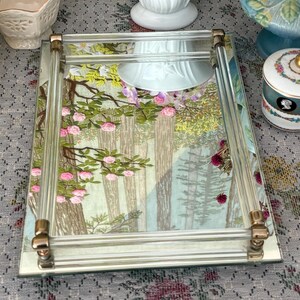 May include: A rectangular mirrored tray with clear acrylic sides and gold-toned corner accents. The tray's mirrored surface features a forest scene with pink roses and green foliage. Decorative item.