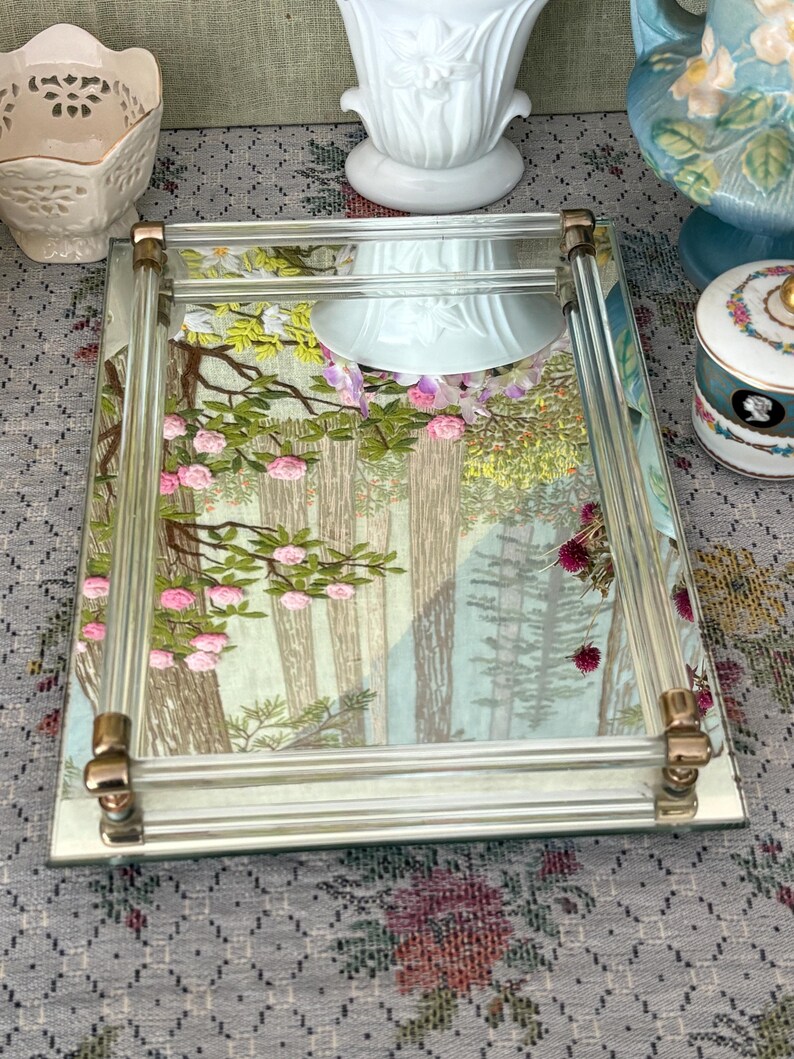 May include: A rectangular mirrored tray with clear acrylic sides and gold-toned corner accents. The tray's mirrored surface features a forest scene with pink roses and green foliage. Decorative item.