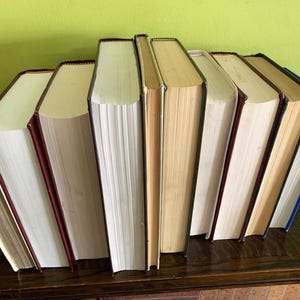 Op de afbeelding: Een verzameling hardcover boeken met verschillende rugkleuren, waaronder rood, blauw en bruin. De boeken zijn gerangschikt op een donkerhouten oppervlak tegen een groene achtergrond. De pagina's zijn gebroken wit.