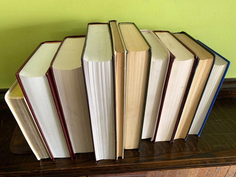 Op de afbeelding: Een verzameling hardcover boeken met verschillende rugkleuren, waaronder rood, blauw en bruin. De boeken zijn gerangschikt op een donkerhouten oppervlak tegen een groene achtergrond. De pagina's zijn gebroken wit.