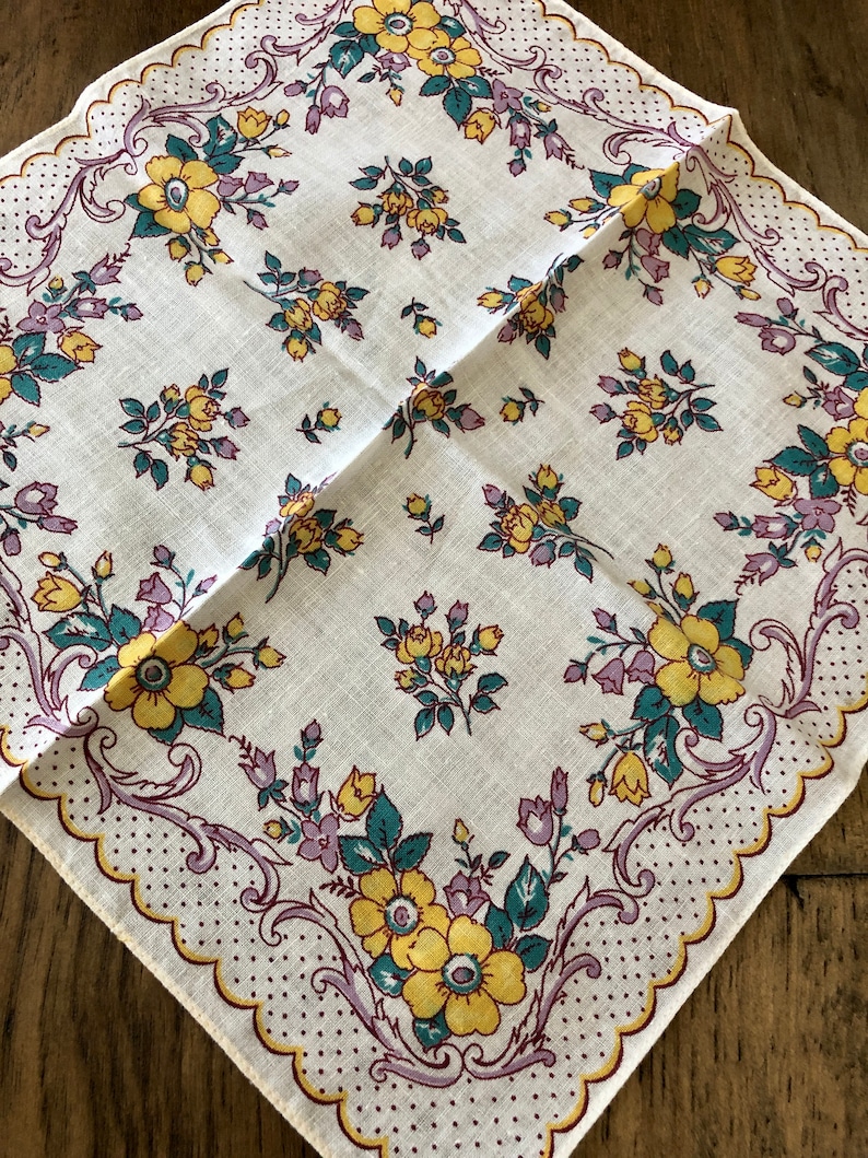 K&ouml;nnte beinhalten: Quadratisches wei&szlig;es Taschentuch mit floralem Muster. Das Design zeigt gelbe und lila Blumen mit gr&uuml;nen Bl&auml;ttern, umrahmt von einem gewellten Rand mit einem gepunkteten Muster. Das Taschentuch ist diagonal gefaltet.