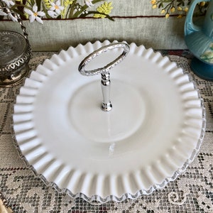 Puede incluir: Un soporte para pasteles de vidrio de leche blanco con un asa plateada. El soporte tiene un borde festoneado y es perfecto para servir postres o aperitivos.