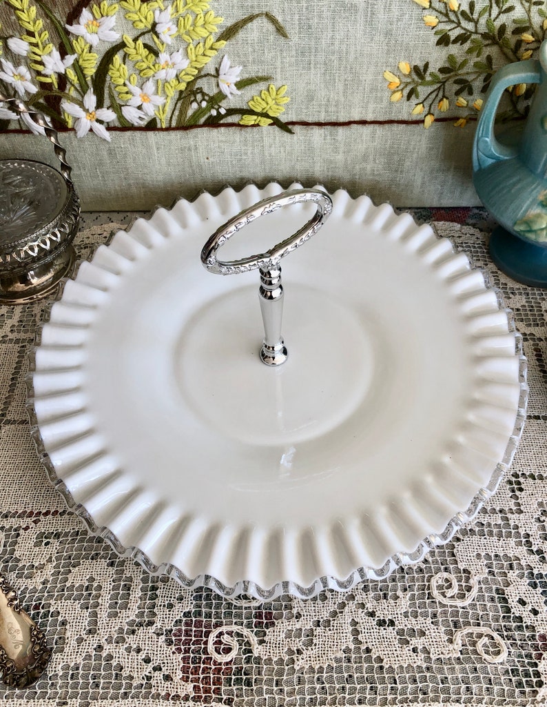 Puede incluir: Un soporte para pasteles de vidrio de leche blanco con un asa plateada. El soporte tiene un borde festoneado y es perfecto para servir postres o aperitivos.