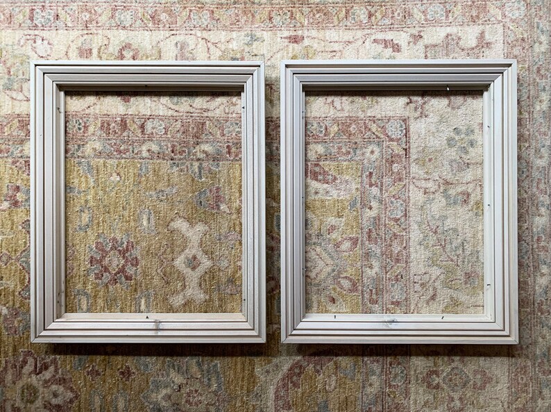 May include: Two white wooden picture frames, each with a simple, rectangular design. The frames are placed side-by-side against a patterned rug.