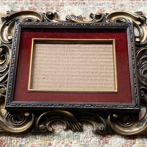 May include: A gold ornate picture frame with a red mat and a blank cardboard insert. The frame has a detailed, intricate design.