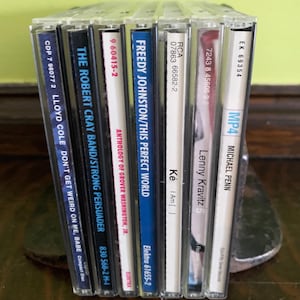 May include: A collection of eight compact discs, including albums by Lloyd Cole, Robert Cray Band, Freddy Johnston, Grover Washington Jr., Lenny Kravitz, and Michael Penn. The CDs are arranged vertically, with the spines facing the viewer. The titles of the albums are visible on the spines.