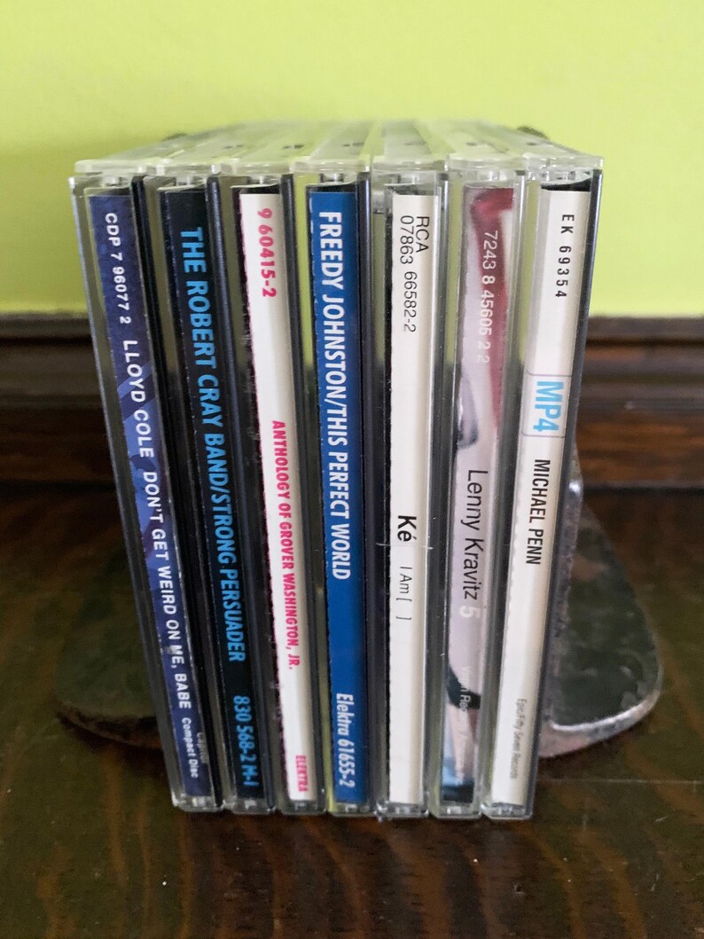 May include: A collection of eight compact discs, including albums by Lloyd Cole, Robert Cray Band, Freddy Johnston, Grover Washington Jr., Lenny Kravitz, and Michael Penn. The CDs are arranged vertically, with the spines facing the viewer. The titles of the albums are visible on the spines.