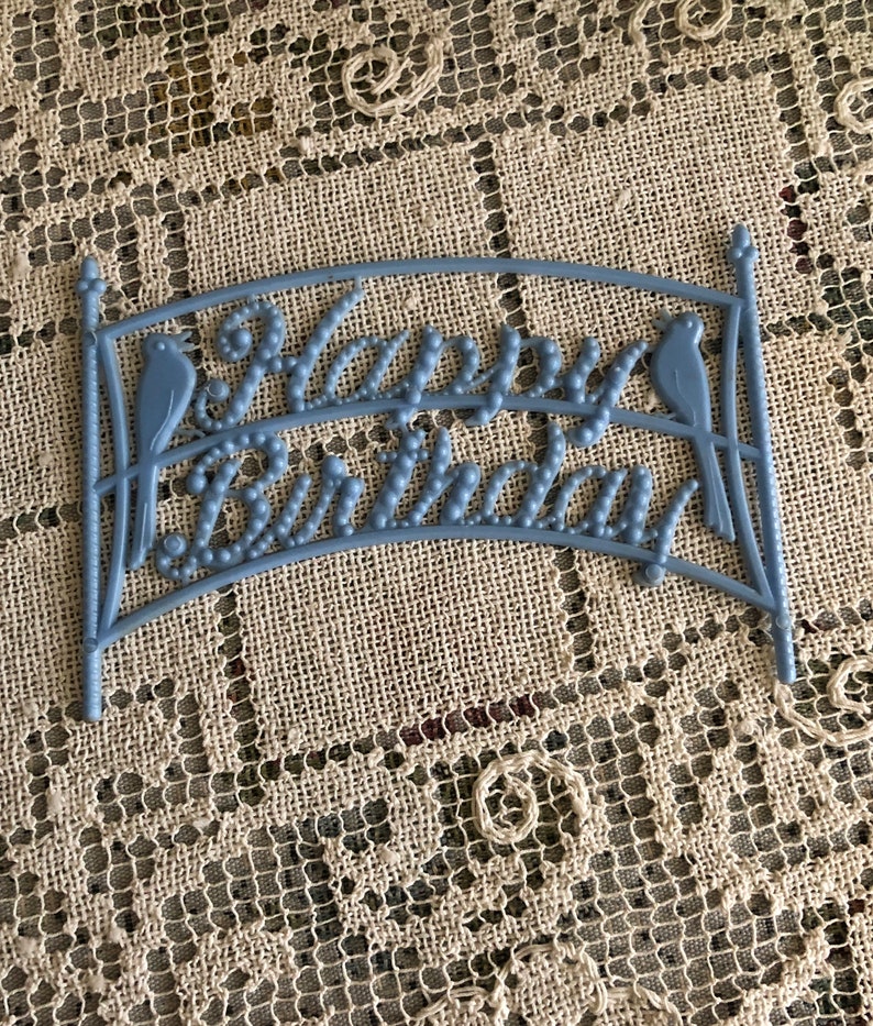 May include: Blue plastic cake topper with the words "Happy Birthday" and two birds on either side.
