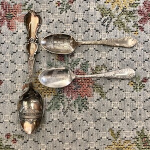 May include: Three silver souvenir spoons with ornate handles and detailed bowls. Two spoons feature engraved buildings, while the third has the word "SOUVENIR" on the handle and a building image. The spoons rest on a floral patterned fabric.