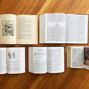 Op de afbeelding: Een bovenaanzicht van vijf open boeken met tekst en illustraties, gerangschikt op een houten oppervlak. De pagina's zijn cr&egrave;mekleurig en de kaften zijn zwart. E&eacute;n boek heeft een bladwijzer met een kleurenfoto.
