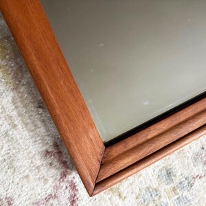 May include: A rectangular mirror with a wooden frame. The frame is a warm brown color and has a grooved design. The mirror's surface reflects the surrounding environment. The mirror is placed on a patterned rug.