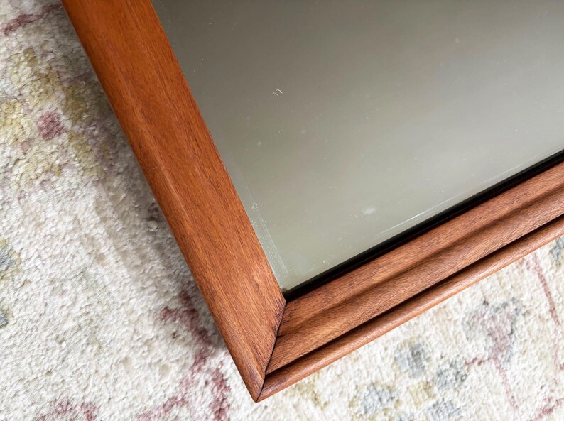 May include: A rectangular mirror with a wooden frame. The frame is a warm brown color and has a grooved design. The mirror's surface reflects the surrounding environment. The mirror is placed on a patterned rug.