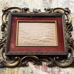 May include: A gold ornate picture frame with a red mat and a brown paper insert. The frame has a detailed, intricate design.