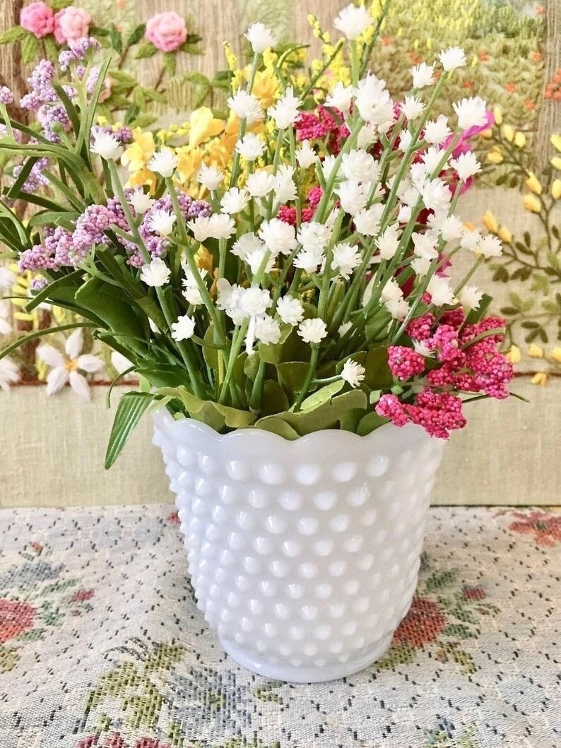 May include: A white hobnail vase filled with a variety of colorful artificial flowers. The flowers include white, pink, yellow, and purple blooms with green foliage. The vase sits on a floral patterned tablecloth.
