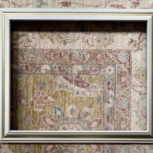 May include: A white picture frame with a gold trim. The frame is sitting on a patterned rug.