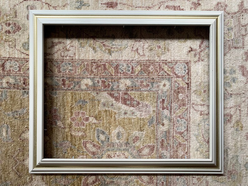 May include: A white picture frame with a gold trim. The frame is sitting on a patterned rug.