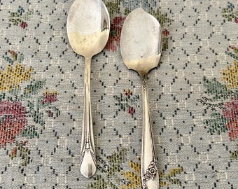 May include: Two silver-toned metal spoons with asymmetrical, leaf-shaped bowls. Each spoon features a slender handle with embossed floral and vine detailing at the base. The metal surfaces show a polished finish with subtle signs of age and wear.