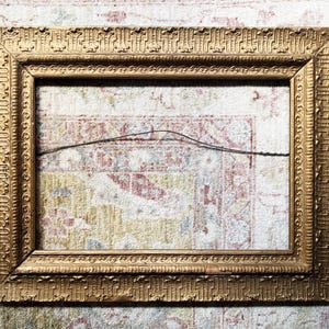 May include: A rectangular, ornate gold picture frame with detailed relief patterns. The frame is empty, with a black wire strung across the back. The frame is set against a patterned rug.