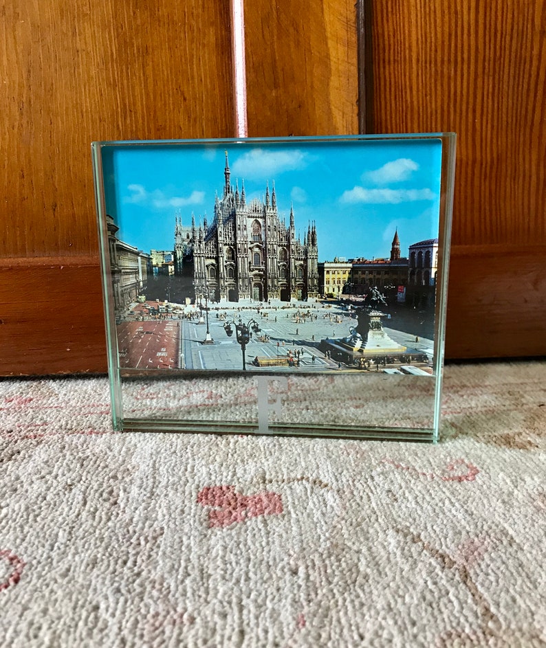 May include: A glass picture frame with a photo of the Duomo di Milano, a large cathedral in Milan, Italy. The photo shows the front facade of the cathedral and the square in front of it.