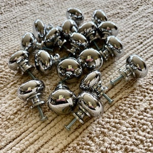 May include: A collection of 18 silver-toned round cabinet knobs with a smooth, shiny finish. The knobs are arranged in a random pattern on a beige textured surface.