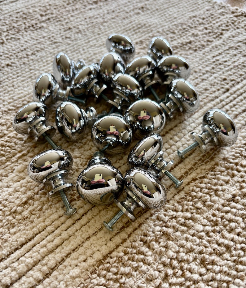 May include: A collection of 18 silver-toned round cabinet knobs with a smooth, shiny finish. The knobs are arranged in a random pattern on a beige textured surface.