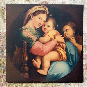 May include: A vintage print of a painting depicting the Madonna and Child with a young angel. The Madonna is wearing a blue and green robe and holding the baby Jesus in her arms. The angel is standing behind them with its hands clasped in prayer.