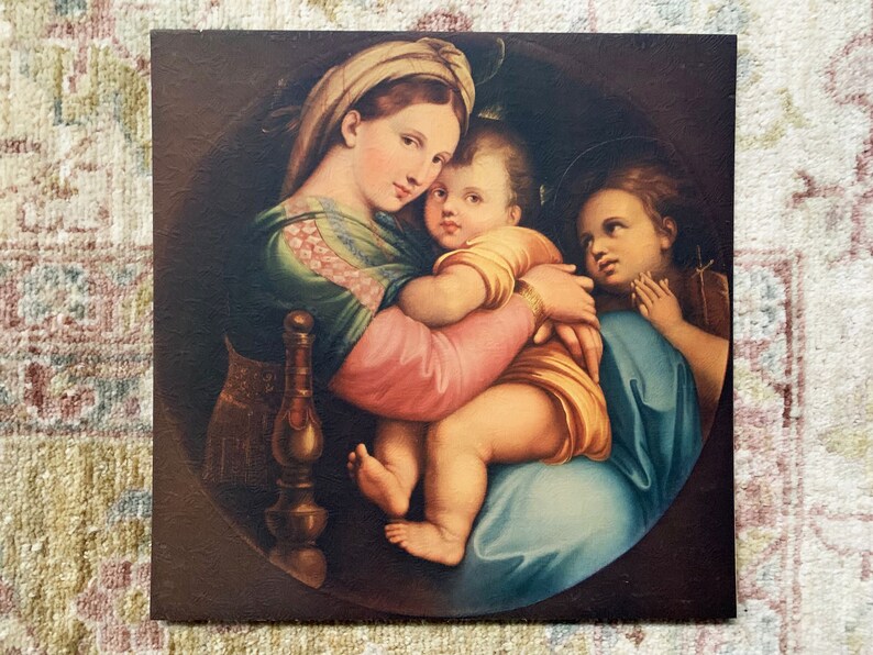 May include: A vintage print of a painting depicting the Madonna and Child with a young angel. The Madonna is wearing a blue and green robe and holding the baby Jesus in her arms. The angel is standing behind them with its hands clasped in prayer.