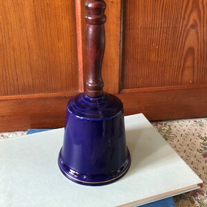 May include: A cobalt blue ceramic bell with a wooden handle. The bell has a gold trim around the rim.