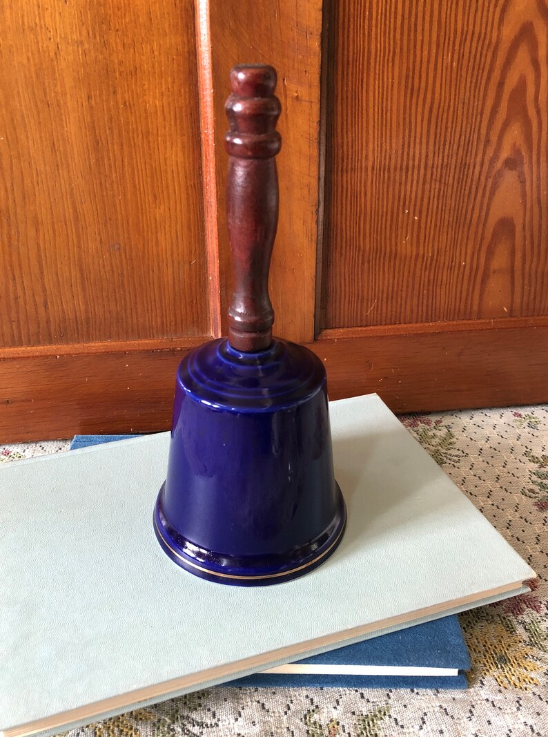 May include: A cobalt blue ceramic bell with a wooden handle. The bell has a gold trim around the rim.