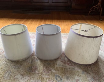 May include: Three white lampshades of varying sizes. The lampshades are made of a fabric material with a subtle texture. The largest lampshade has a light purple trim around the top edge.