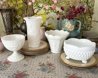 May include: A collection of white milk glass vases and bowls, including a pedestal bowl, a vase with a starburst pattern, and a bowl with a diamond pattern. The items are on a floral patterned tablecloth.