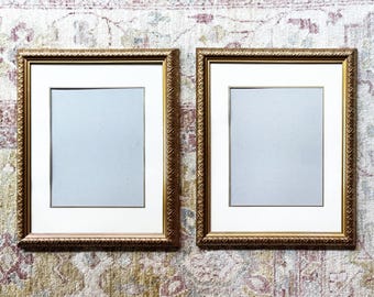 May include: Two rectangular picture frames with ornate gold-toned borders and textured edges. Each frame features a wide white mat board surrounding a light gray rectangular center panel, providing a clean display area for photographs or artwork.