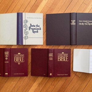 Op de afbeelding: Een assortiment boeken, waaronder Bijbels en religieuze teksten, wordt getoond op een houten oppervlak. De boeken vari&euml;ren in kleur, met sommige met bordeauxrode covers en andere in wit en donkerbruin. Titels zijn onder meer "Into the Promised Land" en "Good News Bible."