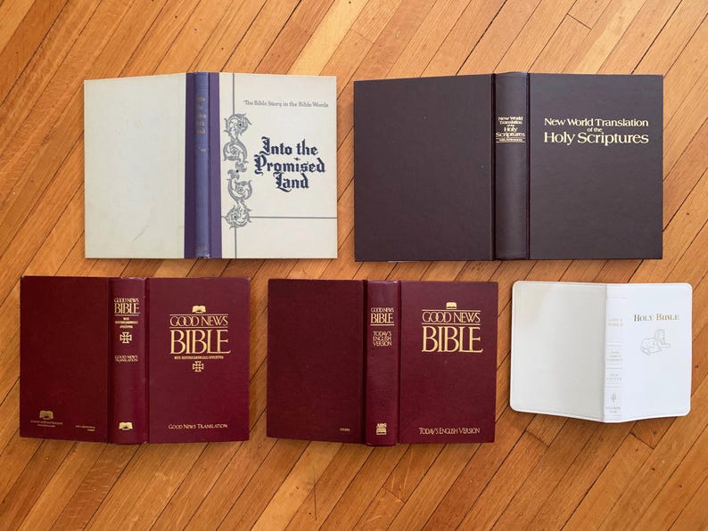 Op de afbeelding: Een assortiment boeken, waaronder Bijbels en religieuze teksten, wordt getoond op een houten oppervlak. De boeken vari&euml;ren in kleur, met sommige met bordeauxrode covers en andere in wit en donkerbruin. Titels zijn onder meer "Into the Promised Land" en "Good News Bible."