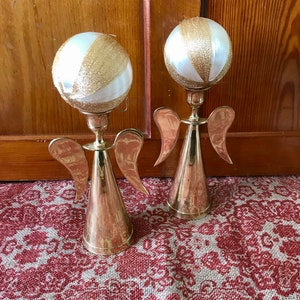May include: Two gold angel candle holders with white and gold ornaments on top. The candle holders are on a red and white patterned surface.