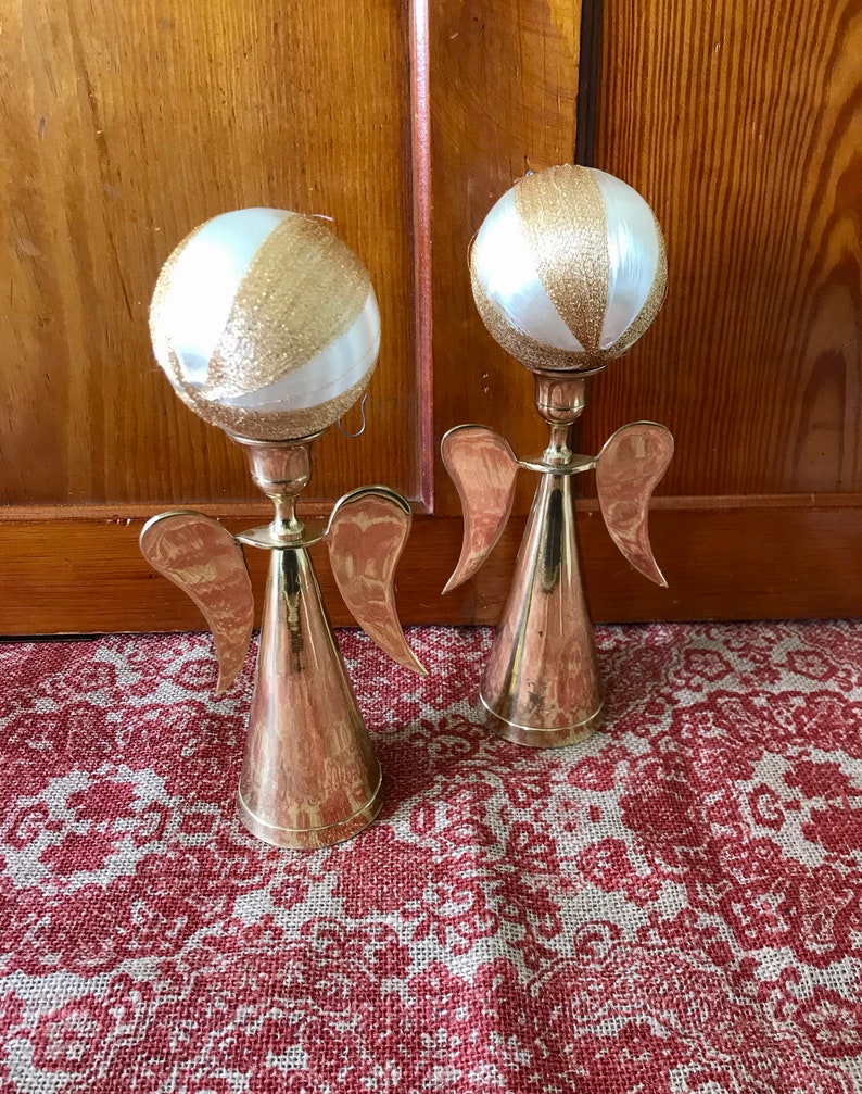 May include: Two gold angel candle holders with white and gold ornaments on top. The candle holders are on a red and white patterned surface.
