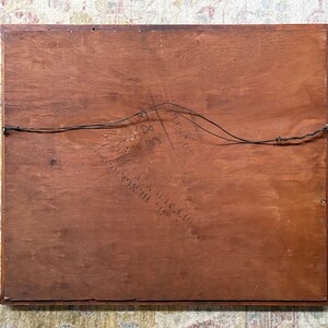 May include: The back of a framed artwork, showing a reddish-brown wooden panel. The wood grain is visible, and a wire is strung across the back for hanging. Some text is visible, but it is difficult to read.