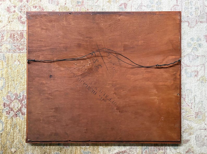 May include: The back of a framed artwork, showing a reddish-brown wooden panel. The wood grain is visible, and a wire is strung across the back for hanging. Some text is visible, but it is difficult to read.