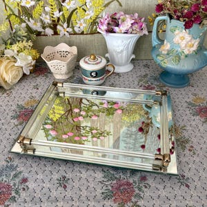 May include: A rectangular mirrored tray with gold-toned accents, reflecting a floral tapestry. The tray is surrounded by decorative vases and a teacup, with a floral arrangement in each. The scene is set on a floral patterned tablecloth.