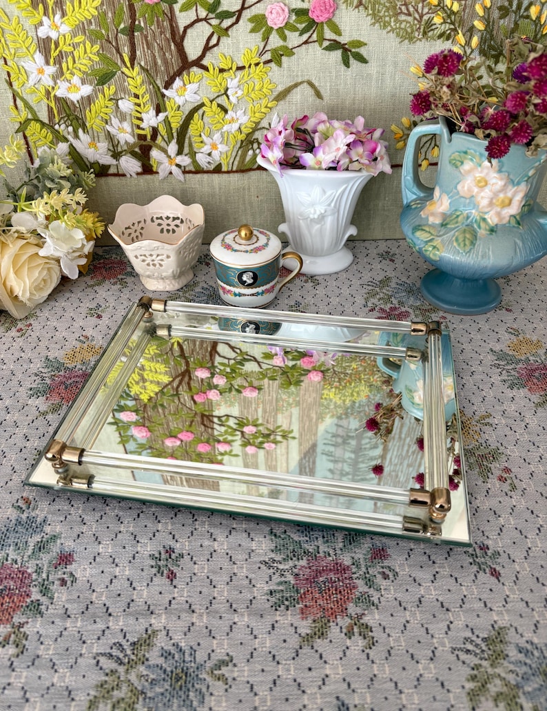 May include: A rectangular mirrored tray with gold-toned accents, reflecting a floral tapestry. The tray is surrounded by decorative vases and a teacup, with a floral arrangement in each. The scene is set on a floral patterned tablecloth.