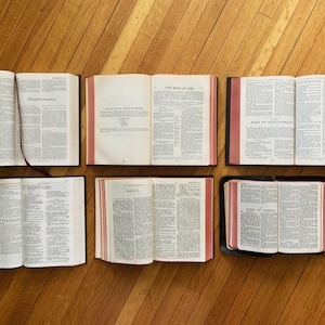 May include: Six open books with red and black covers, laid out on a wooden floor. The books are open to different pages, showing text in black and white. The books are titled "Deuteronomy", "The Book of Job", "Book of Ezekiel", "Samuel", and "Book of Ezra".