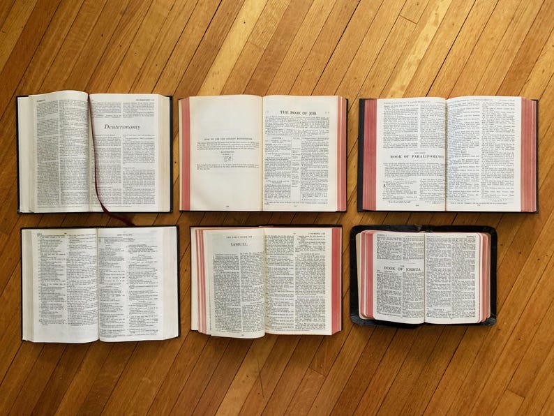 May include: Six open books with red and black covers, laid out on a wooden floor. The books are open to different pages, showing text in black and white. The books are titled "Deuteronomy", "The Book of Job", "Book of Ezekiel", "Samuel", and "Book of Ezra".